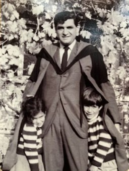 A black and white photo of a young Peter in a suit with a graduation gown. Two children in striped shirts hide underneath.