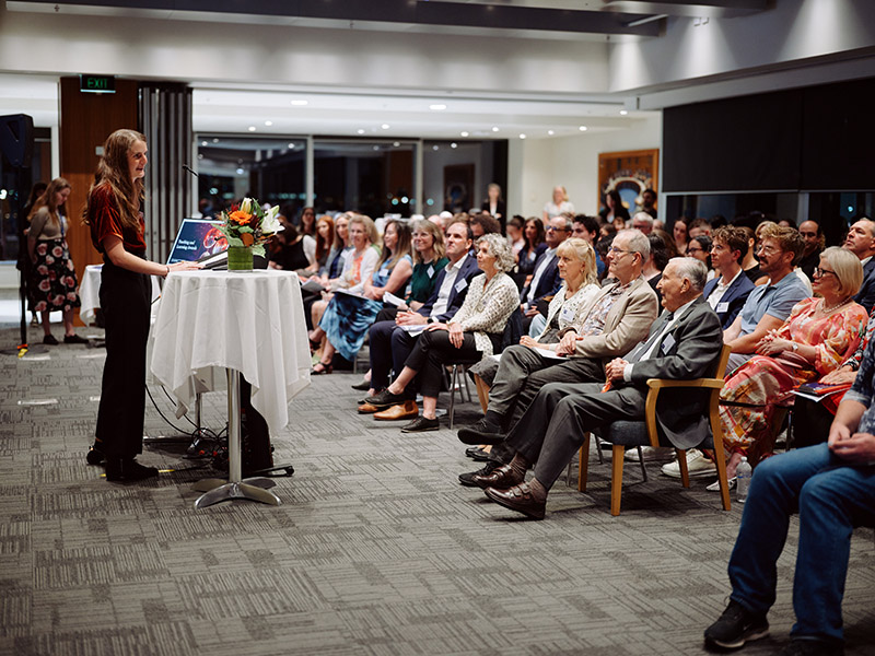 MC Dr Elise Kalokerinos presents at a podium, with a full audience of prize winners and guests watching.