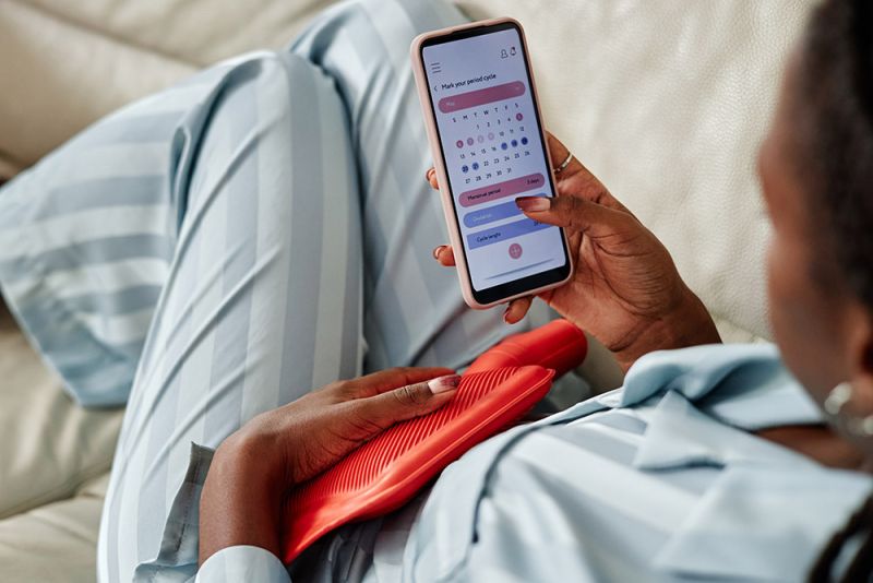 A woman sits in pyjamas with a hot water bottle on her stomach. In her hand, she's looking at a period tracker on her phone.