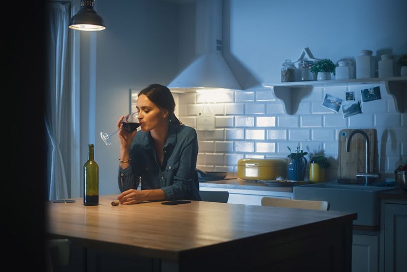 A woman sits alone in a kitchen, drinking a glass of wine.