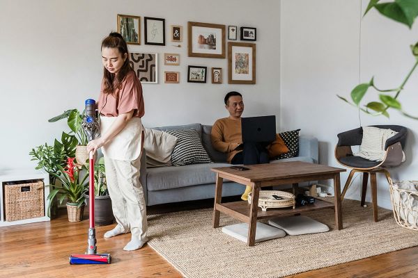 A woman vacuums the floor, while a man relaxes on the couch behind her with his laptop.