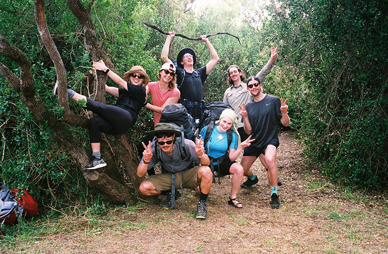A group of young people wearing hiking gear and backpacks make fun poses on a trail.