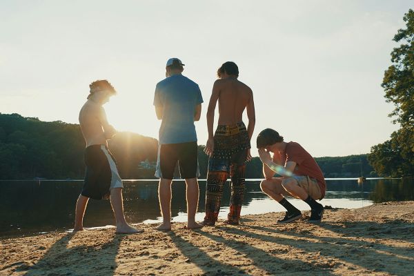 A group of teenage boys stand on the beach looking out over the water.